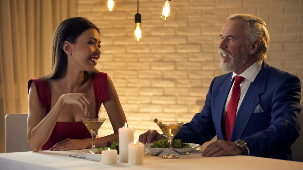 Ein älterer Herr in einem blauen Anzug und einer jungen Dame in einem roten Kleid genießen ein romantisches Abendessen, ein typisches Setting für ein Fetishescort-Dinner-Date.