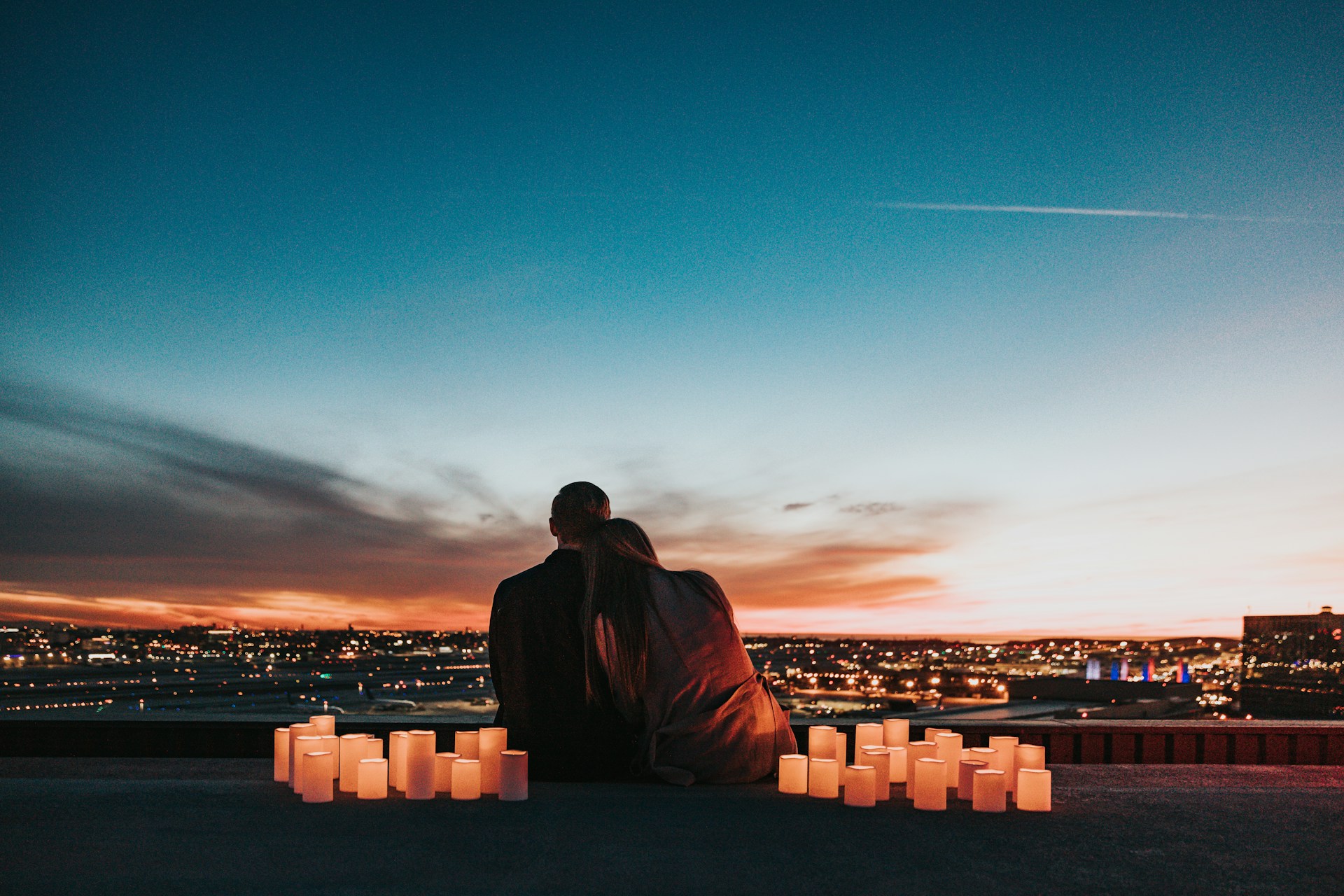 Ein Paar genießt einen romantischen Sonnenuntergang auf einer Dachterrasse, umgeben von Kerzen, mit einem Panoramablick auf die Stadtlichter.